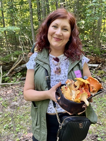 woman in T-shirt and vest holding a baseball cap full of mushrooms while standing in a forested area