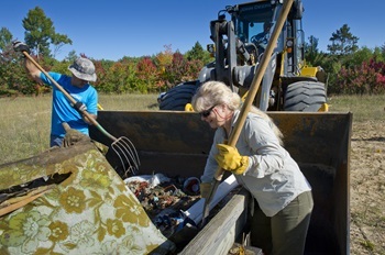 a man and a woman, each wearing gloves and using a pitchfork, load garbage into a dumpster during Adopt-a-Forest cleanup