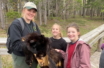 A woman in a DNR baseball camp shows an animal pelt to two young, smiling girls. They are standing outdoors near end of a wooden bridge