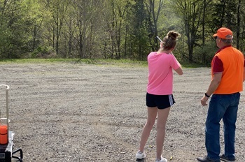 A girl in a T-shirt and shorts holds and aims a gun at a shooting range as a volunteer safety instructor in an orange vest watches