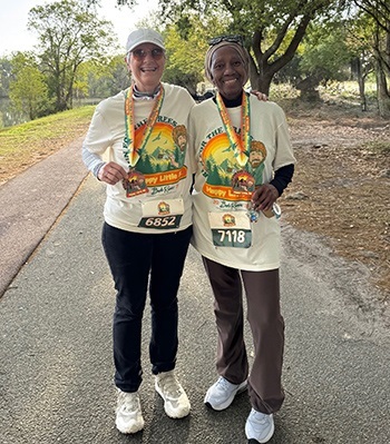 Two women in Happy Little Trees T-shirts, medals and race bibs smile as they stand side by side on a paved park trail