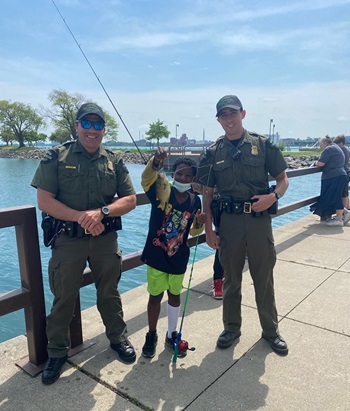 two male DNR conservation officers in uniform stand next to a little boy proudly showing off the fish he caught
