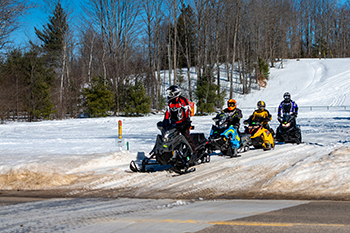 A group of snowmobilers stops at the edge of a road look for traffic before crossing.