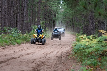 three off-road vehicles move single-file down a wide, dirt path in a mature, thick forested area 