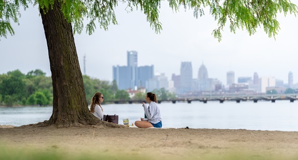 Two women holding drinks sit on the ground under a large, shading tree at Belle Isle Park. Windsor city skyline is fuzzy in background
