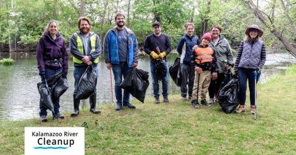 small group of adults and a young boy dressed in outdoor gear hold garbage bags and trash pickup sticks along a riverbank