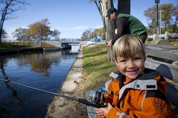 a little blond-haired boy with a big smile and orange jacket holds a fishing pole over the water as a man in green DNR vest fishes behind him