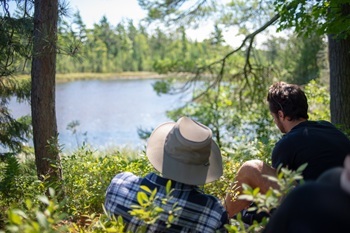 Two people in T-shirts, one in flannel, sit on a forested rise, looking out over the water at Wilderness State Park