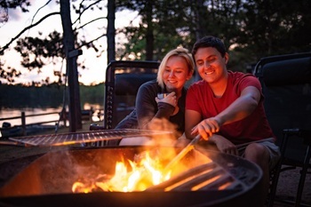 smiling woman and man sitting closely by a campfire as he pokes it with a stick, while dusky evening sets in over the nearby lake