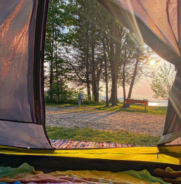 Looking out of a folded-up tent door, with a brightly colored towel on the floor, at sunny Lake Charlevoix. A park bench sits near the shore.