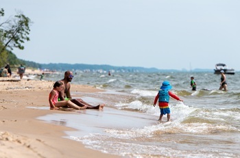 A young child in brightly colored hat, swimsuit and life vest splashes at the lakeshore, as a man and young boy sit nearby