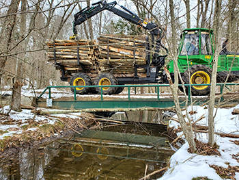 A load of logs is hauled across a bridge over a creek amid a wintry scene.