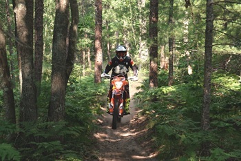 a dirt bike rider wearing a helmet heads up a trail in a thickly forested area on a sunny day
