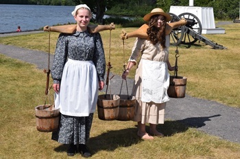 Young girls in Civil War-era clothing carry bucket holders across their shoulders at Fort Wilkins Historic State Park