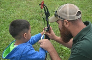a man in a ballcap shows a young boy wearing a hoodie how to properly hold and use an archery bow outdoors