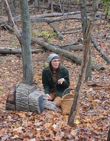 woman in a hat and jacket, holding mushrooms, sits on ground next to tree stumps in a forested area