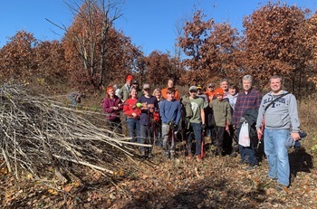 About a dozen people in outdoor clothing hold bags while standing next to a brush pile on a cleanup day at Island State Recreation Area