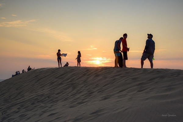 Adults and children atop the sand dunes at Silver Lake State Park are captured in shadow as the sun sets against a golden sky