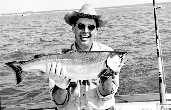 black and white picture of Dr. Howard Tanner, smiling broadly in sunglasses and hat, on a boat holding up a salmon caught on the Great Lakes