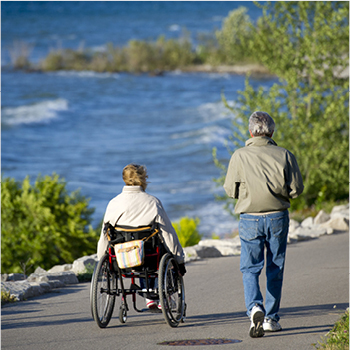 A couple enjoying a trail along a lake. One person is in a wheelchair while someone walks alongside. 