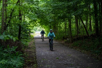 two bicyclists wearing jackets and helmets pedal down a paved, forested stretch of the Kal-Haven Trail in southwest Michigan