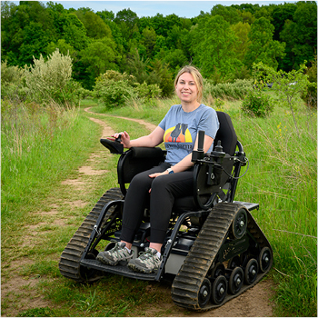 A young woman in a track chair, smiling along a trail