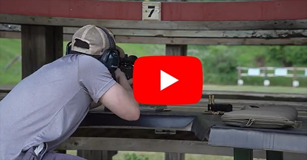 a red video "play" button overlaid on image of a person leaning on a shooting range counter, aiming a rifle at a downrange target