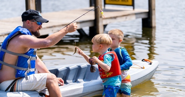A man in a kayak holds a fishing pole with a small fish on the line as two little boys in life jackets stand in shallow water, admiring the catch