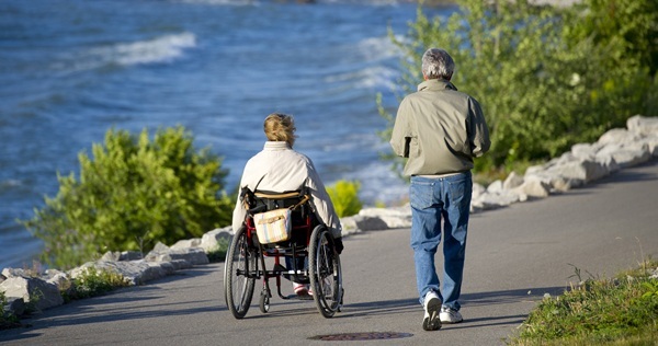 A person walking and a person in a wheelchair, both dressed in fall clothing, go down the paved Little Traverse Bay Wheelway along the water