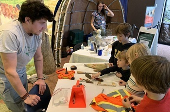 Young children handle and ask questions about some historical tools on a table at Archaeology Day event, as a young man listens