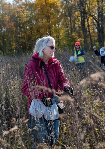 A smiling, older woman wearing sunglasses and a fall coat helps collect native seeds at Fort Custer Recreation Area (autumn 2022).