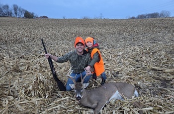 A smiling man and boy pose in a grassy field with a deer from a successful hunt. Both are wearing camo and hunter orange hats.