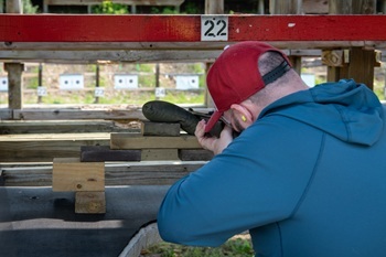 man in a red cap and blue shirt looks toward a target as he aims a rifle, leaning on a counter at a shooting range