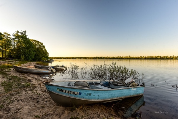 Half a dozen fishing boats, a pale blue one in foreground, sit on the sand near shallow water of Muskallonge Lake, Luce County