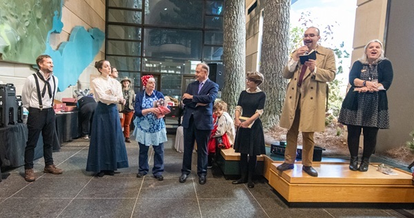 man and woman stand on a raised seating area in museum atrium, addressing a crowd, surrounded by people in historical costumes