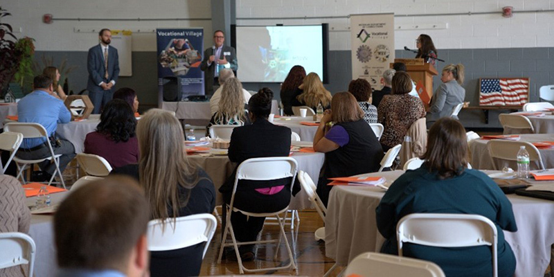 Room full of people seated at tables and chair listening to a speaker