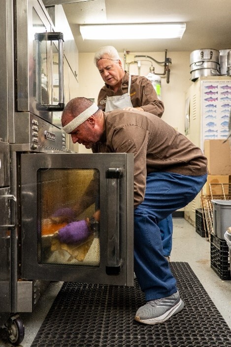 Two men working in a kitchen as one of them places a pan in the oven