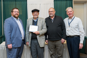 Four men pose for a picture, one man in a hat holds up a framed document.