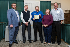 Four men and a woman pose for a picture, one man holds up a framed document.