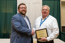 Two men pose for a picture, shaking hands, while one of the men holds up a framed document.