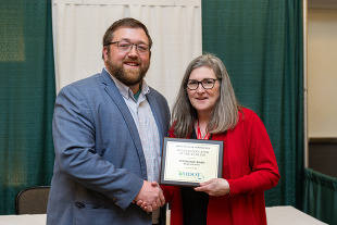 A man and woman pose for a picture, shaking hands, while the woman holds up a framed document.