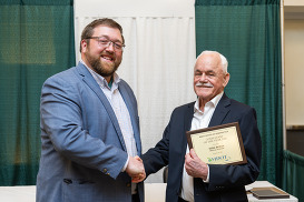Two men pose for a picture, shaking hands, while one of the men holds up a framed document.