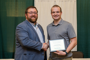 Two men pose for a picture, shaking hands, while one of the men holds up a framed document.
