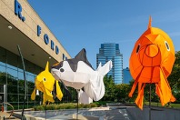 An outdoor art installation featuring colorful fish in yellow, white and orange with buildings in the background.