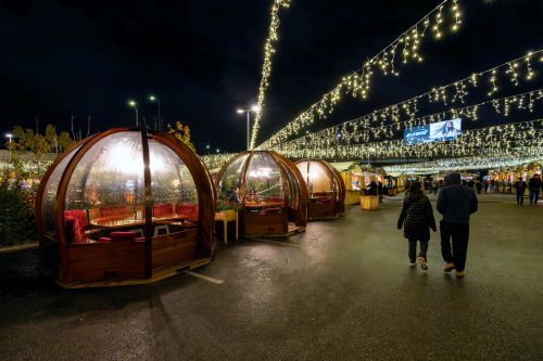 People walking beside heated igloo tents under holiday lights at night.
