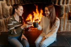 Two women with cocktails beside a fireplace. 