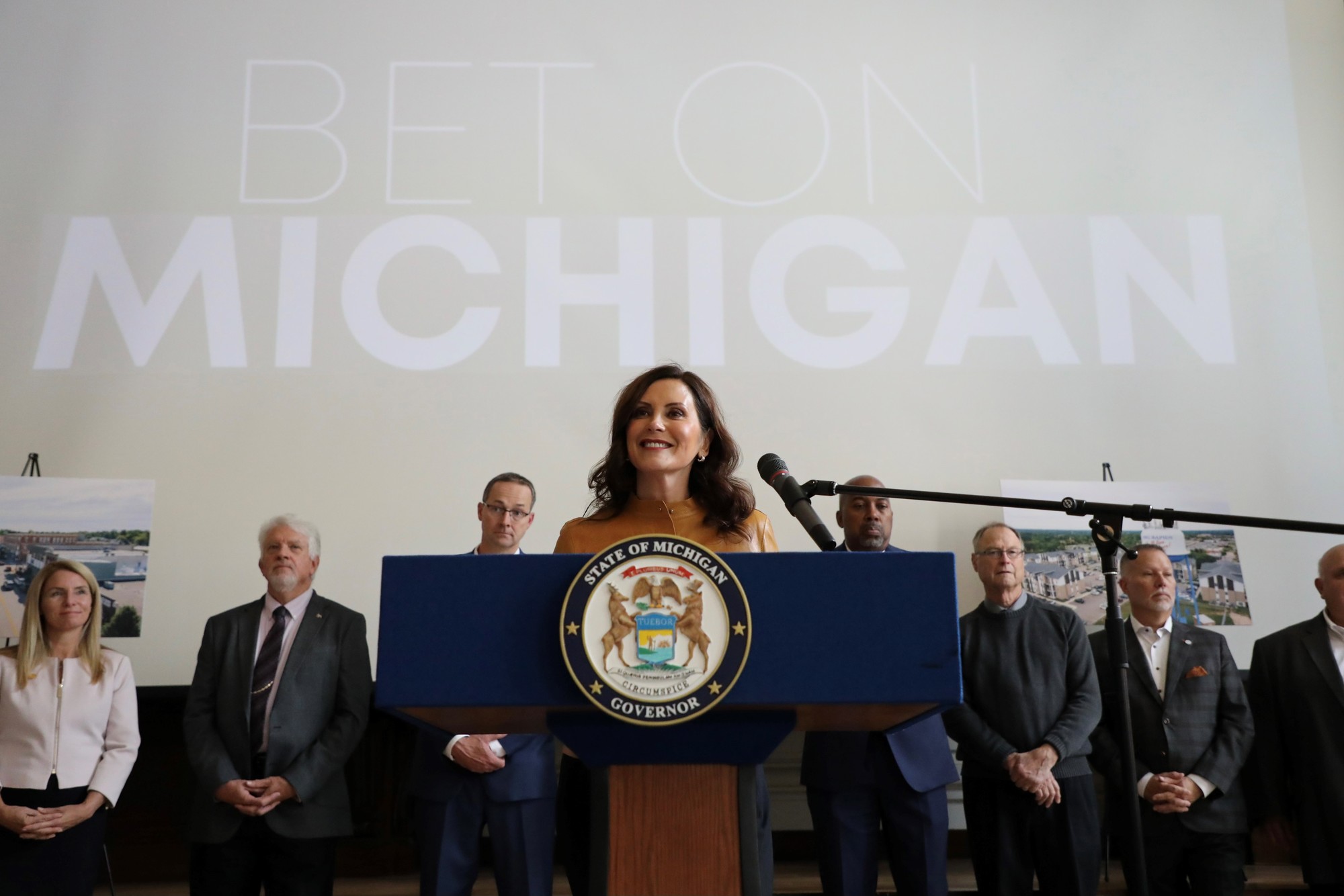 Gov. Whitmer stands at podium at event. Behind her, a screen with the text 'Bet on Michigan' is displayed 