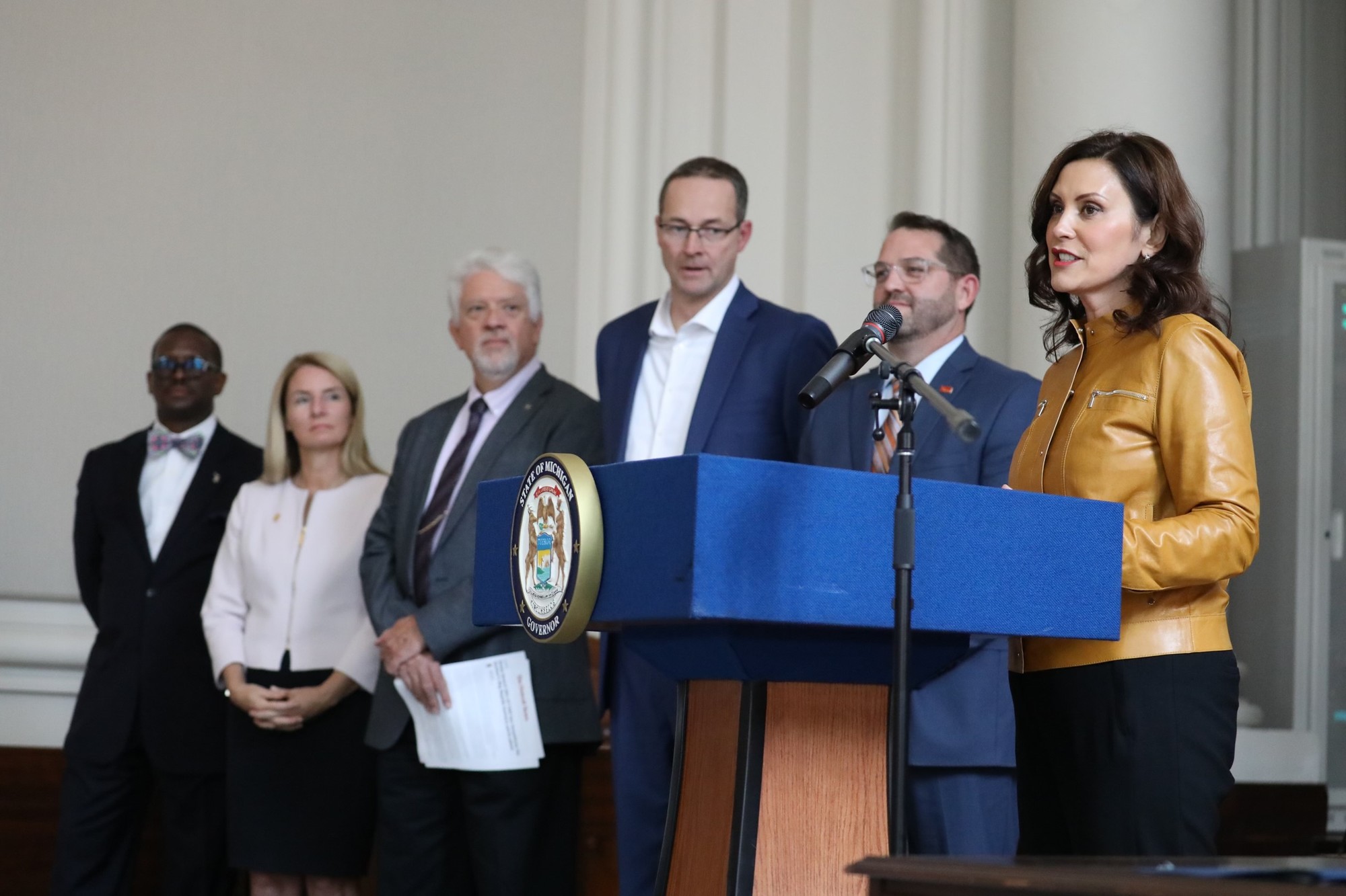 Gov. Whitmer speaks at podium during event. A group of people stand to her right side. 