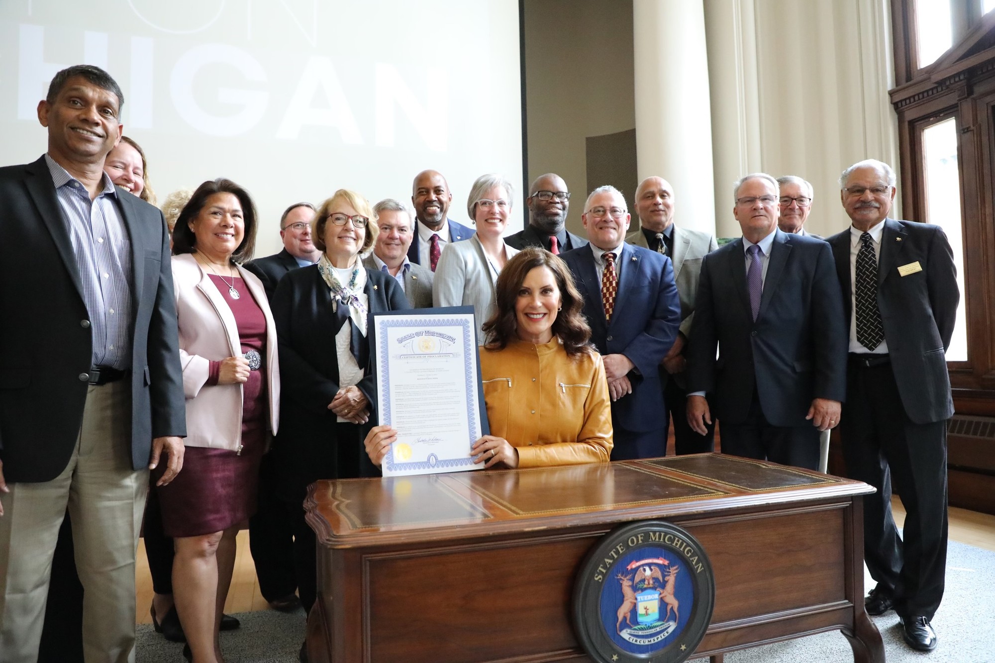 Gov. Whitmer, sitting at desk, holds up the signed bill. There is a group of people behind her. 