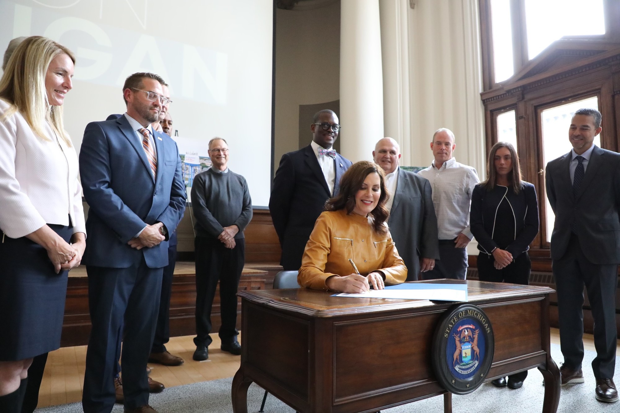 Gov. Whitmer sits at desk, signing bill. There is a group of people behind her watching her sign the bill 